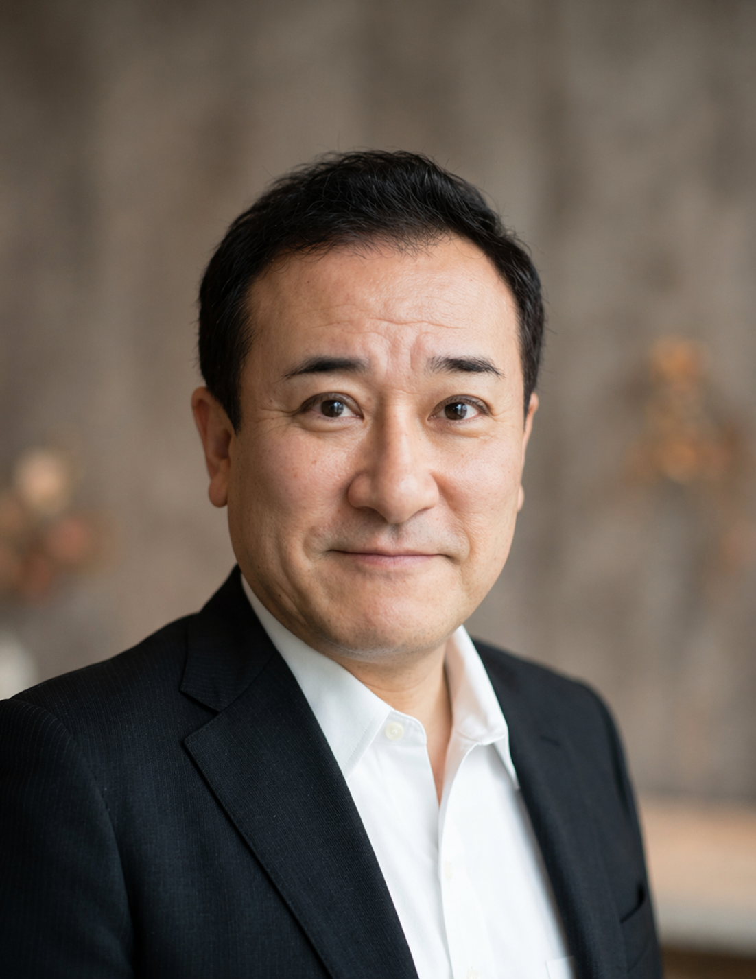 白いシャツと黒いジャケットを着たこにしさんがテーブルに手を置き、笑顔で正面を向いているポートレート写真